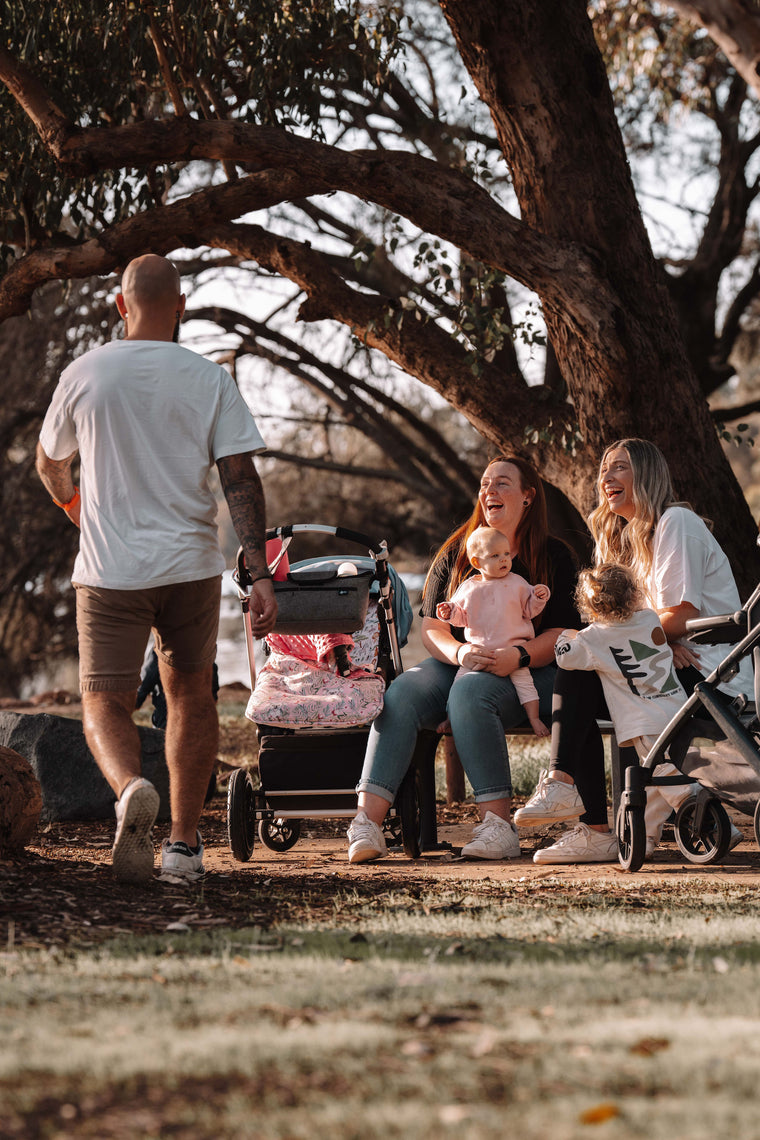 Family with children and strollers sitting under a tree in a park