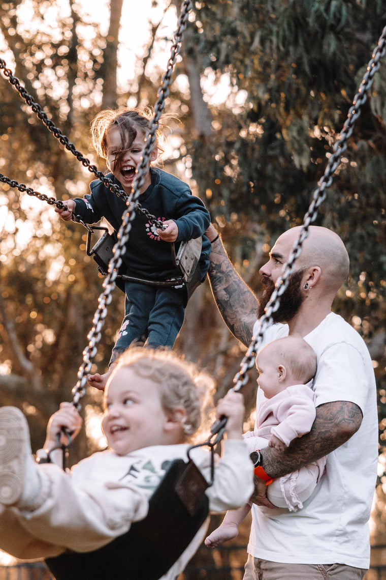 Family of four enjoying a day at the park with children on swings.