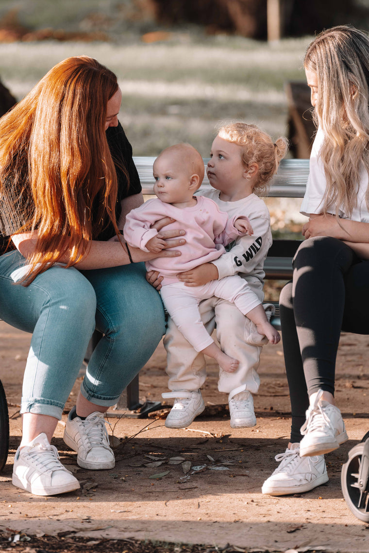 Two women sitting on a wooden bench with two young children, one of whom is holding a baby.